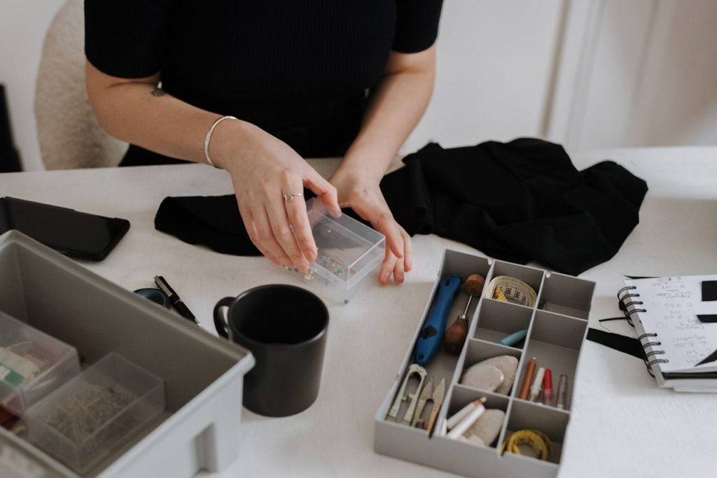 Unrecognizable female dressmaker with box of pins standing at table with containers full of sewing supplies while working in light workshop